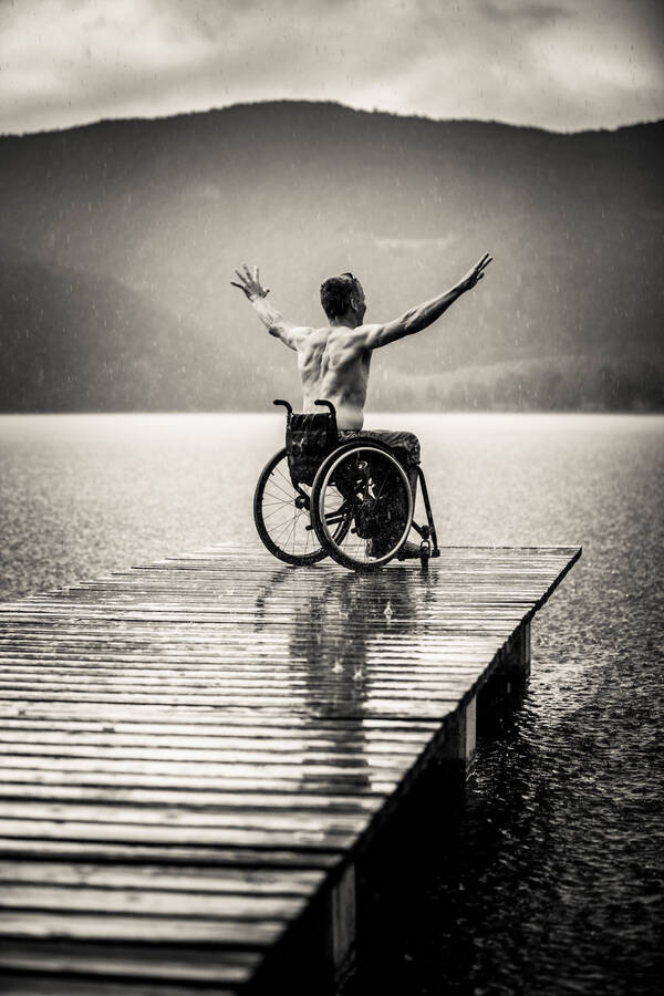 Ein Mann im Rollstuhl auf einem Steg eines Sees. Schwarz-Weiß