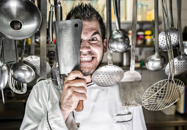 Portrait eines Kochs, der in einer Küche zwischen hängenden Schöpfkellen steht und eine Gesichtshälfte mit einem Fleischerbeil verdeckt und dabei diabolisch Lacht.