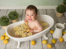 Ein Baby sitht in einer kleinen weißen Badewanne. Im Wasser schwimmen viele Zitronenscheiben, neben der Wanne stehen Kräutertöfpe, weiße Kannen und liegen viele ganze Zitronen 