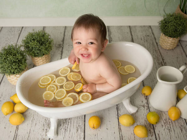 Ein Baby sitht in einer kleinen weißen Badewanne. Im Wasser schwimmen viele Zitronenscheiben, neben der Wanne stehen Kräutertöfpe, weiße Kannen und liegen viele ganze Zitronen 