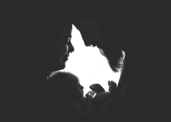 Ein bärtiger Mann und eine Frau (beide mit Nasenpiercing) bilden gemeinsam mit einem neugeborenen Baby einen Silhouetten-artigen Rahmen. Schwarz-Weiß.