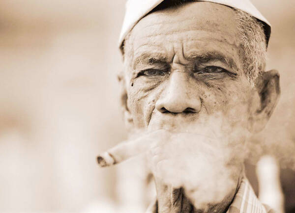Portrait eines alten, dunkelhäutigen Mannes mit einer weißen Kopfbedeckung, der eine Zigarre raucht. Sepiatönung.