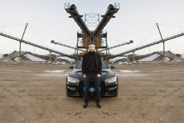 Foto von einem Mann, der zentral im Bild vor einem Auto steht, im Hintergrund diverse Industrieanlagen. Das gesamte Bild ist bis auf den Mann in der Mittenachse horizontal gespiegelt.