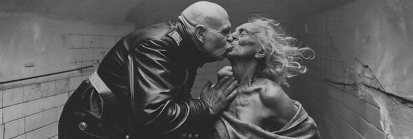 In einem weiß gefliesten, baufälligen niedrigen Raum Küsst ein alter, glatzköpfiger Mann in einem schwarzen, militärischen Ledermantel stürmisch eine alte weißhaarige Frau, die nur in eine Decke gehüllt ist. Schwarz-Weiß.