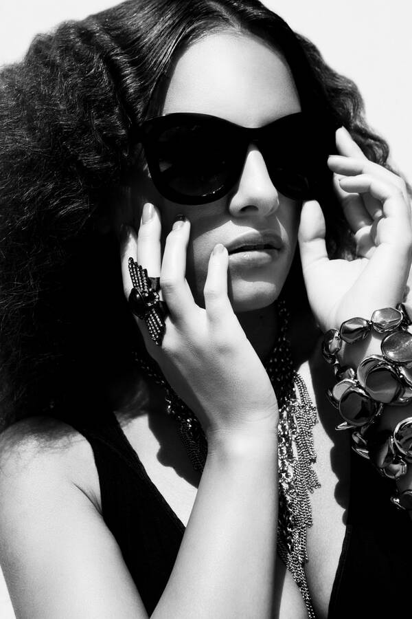 Portrait einer jungen Frau mit großer Sonnenbrille, beide Hände beim Gesicht. Schwarz-Weiß.