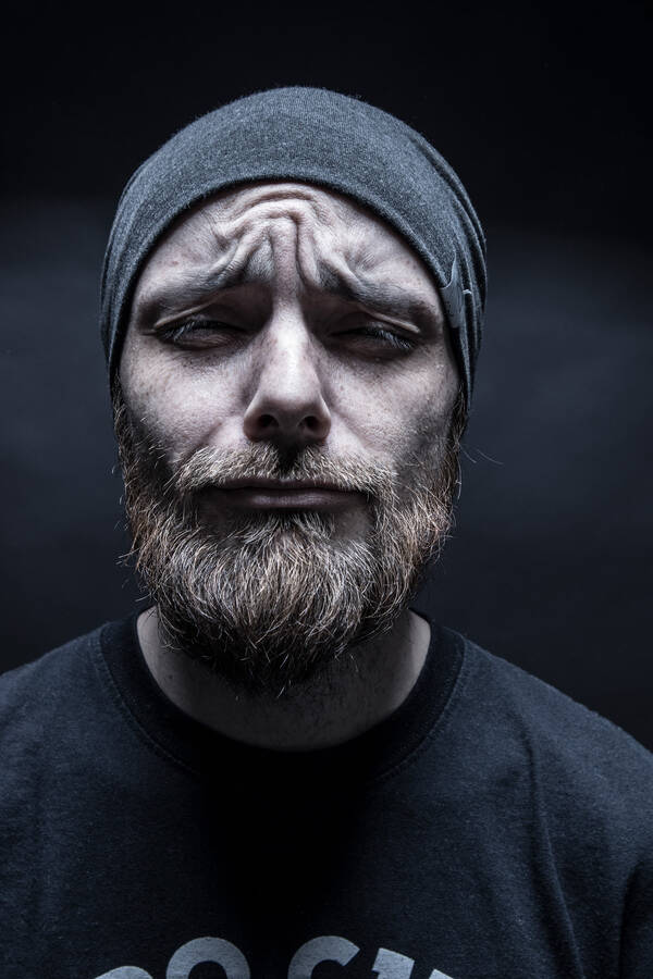Portrait eines bärtigen Mannes mit geschlossenen Augen, die Stirn in Falten gelegt. Der Mann trägt eine graue Mütze.