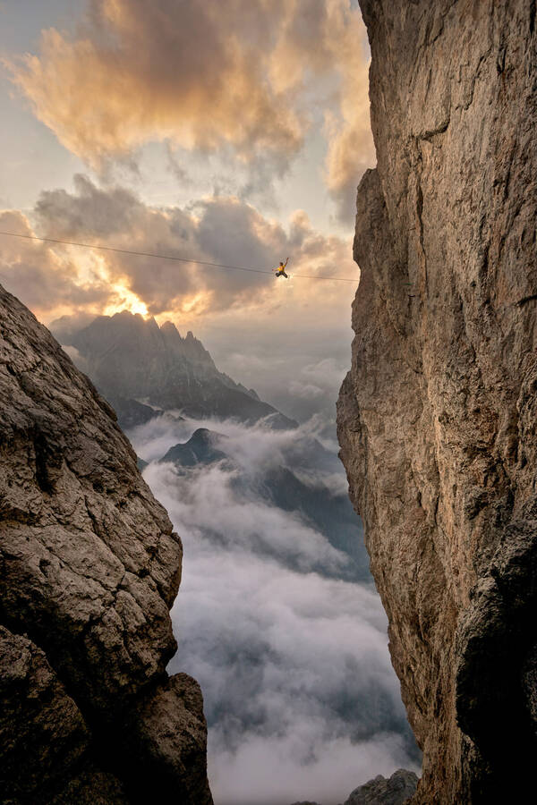 Ein Athlet überwindet einen Felsabgrund im Gebirge auf einer Slackline. Im Hintergrund werden felsige Berge spektakulär in Wolken und Nebelschwaden gehüllt.