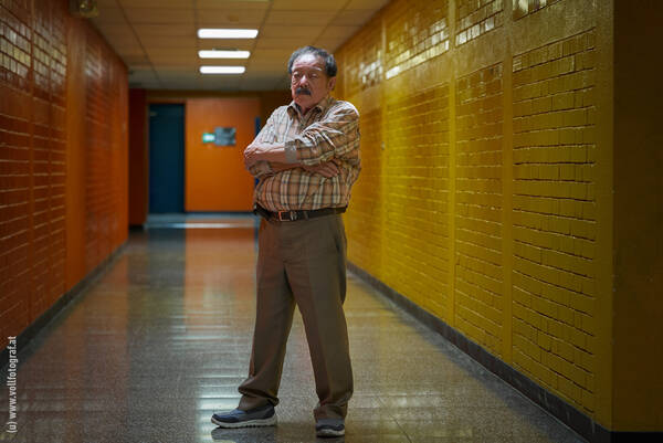 Ein älterer Mann steht mit verschränkten Armen in einem gelb gefliesten Gang mit orangen Kabinentüren.