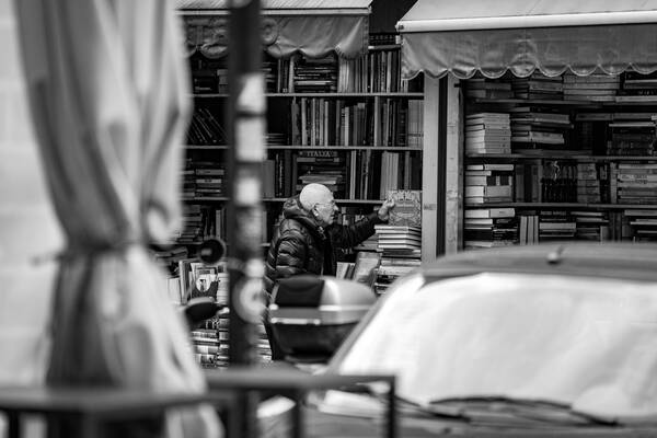 Blick über Fahrzeuge auf einer Straße in ein zur Straße hin offenes Antiquariat. Ein Mann hat ein Buch mit dem Titel 