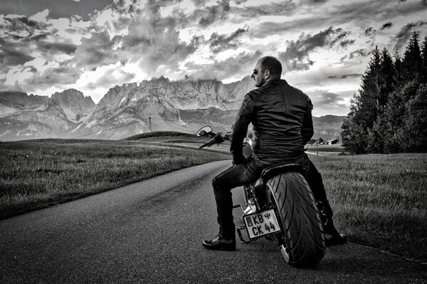 Ein Mann mit Sonnenbrille sitzt auf einem Motorrad und schaut in die Ferne. Im Hintergrund ist der Gebirgszug des Wilden Kaisers zu sehen. Schwarz-Weiß.