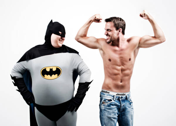 Ein Korpulenter Mann in einem Batmankostüm und ein Mann mit naktem Oberkörpber der eine Krafttrainingspose, schauen sich an