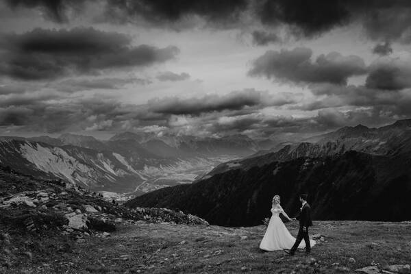 Ein Brautpaar geht hand in Hand über eine Bergwiese mit Blik ins Tal und dramatischer Wolkenstimmung. Schwarz-Weiß.
