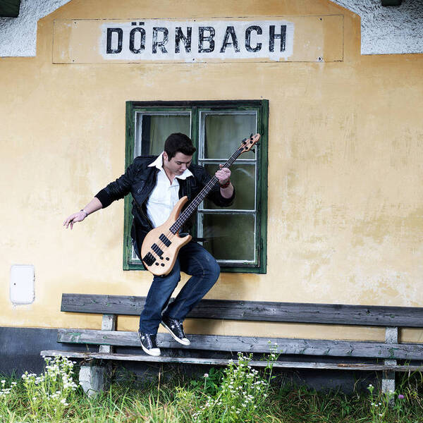Ein Musiker mit Bassgitarre steht auf einer Bank vor dem Bahnhof Dörnbach.