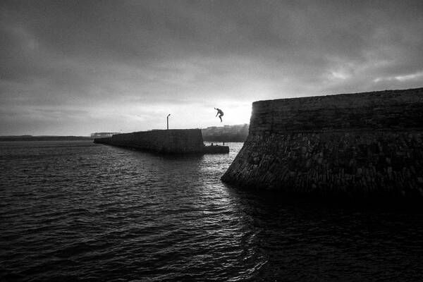 Ein Mensch springt im Gegenlicht von der Hafenmauer ins Meer. Schwarz-Weiß