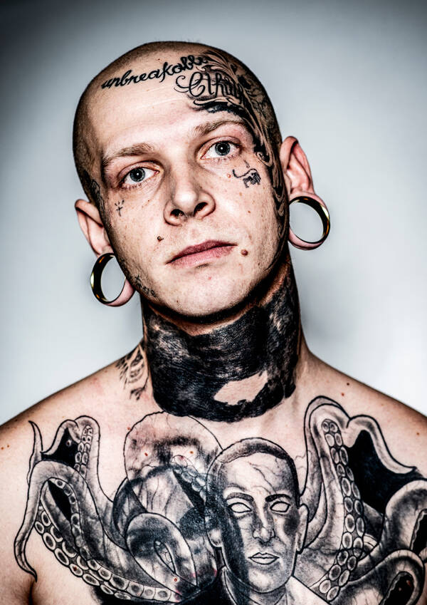 Portrait eines jungen Mannes, dessen Brust, Hals, Gesicht und kahlrasierter Schädel mit schwarzen Bildern tätowiert sind. Auf dem Kopf steht 
