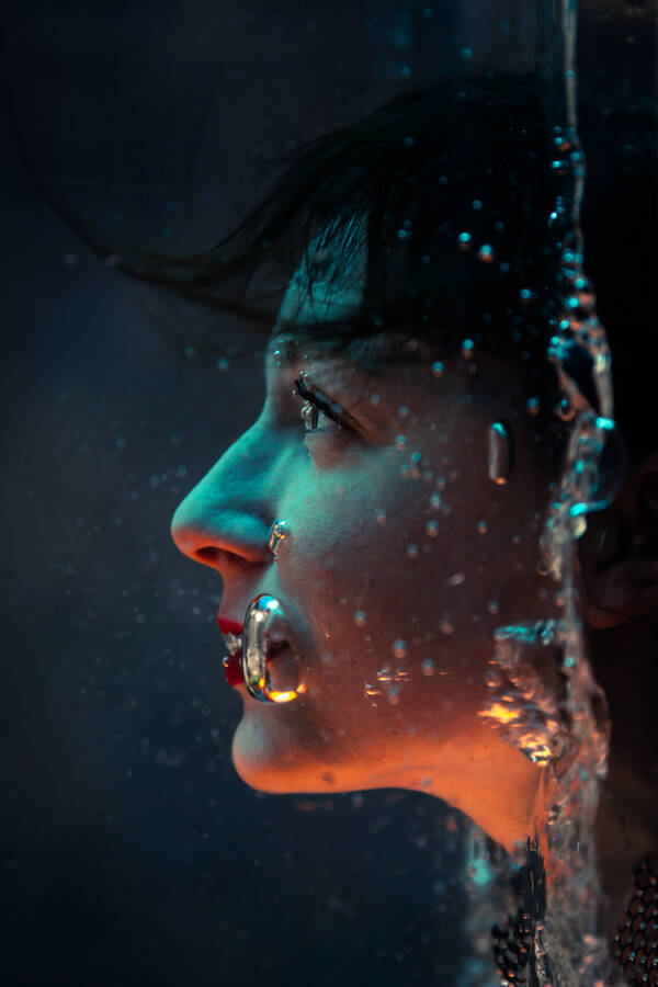 Eine Frau hält ihr Gesicht unter Wasser, das Bild ist dabei um 90° im Uhrzeigersinn verdreht, so dass die Wasserlinie am rechten Bildrand von oben nach unten verläuft.