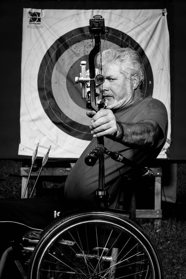 Ein älterer, weißhaariger, bärtiger Mann in einem Rollstuhl hält einen Sportbogen und Pfeile. Im Hintergrund sieht man eine Zielscheibe. Schwarz-Weiß.