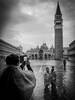 Venedigs Markusplatz mit dem Campanile bei Hochwasser. im Vordergrund macht ein Tourist ein Foto mit dem Mobiltelefon von einem asiatischen Paar, das im Wasser steht. Im Hintergrund steht ein Mann mit Stativ und Regenschirm im Wasser um zu Fotografieren.