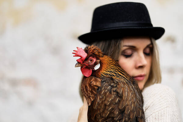 Eine Frau mit schwarzem Hut hält ein braunes Huhn an ihre Schulter gedrückt.