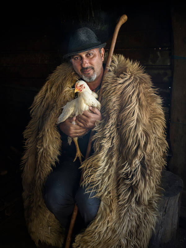 ein Hirte mit Filzhut und in Fell gekleidet hält ein Huhn in Händen.