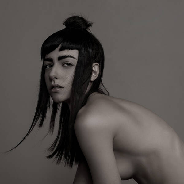 Portrait einer unbekleideten jungen Frau mit langen, teils zu einem Knoten hochgebundenen dunklen Haaren. Die Frau ist vornübergebeugt.