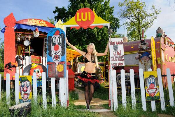 Eine spärlich bekleidete Frau steht im Tor eines Lattenzaunes vor bunt bemalten Wohnwägen. Bilder von Clowns sind auf den Zaun gehängt.