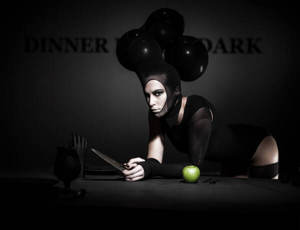 Frau in ausgefallenem schwarzen Designoutfit lehnt mit einem großen Messer auf einem Speisetisch mit einem grünen Apfel.