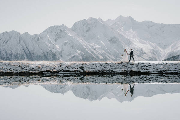 Ein Brautpaar geht vor verschneiter Bergkulisse. Die Berge und das Brautpaar spiegeln sich im Speicherteich.