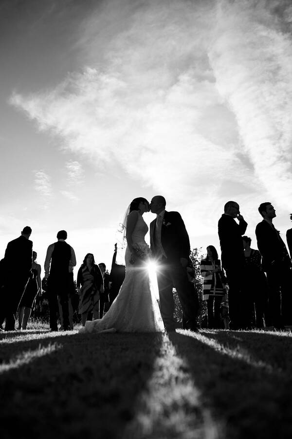 Silhouette eines küssenden Brautpaares inmitten der Hochzeitsgäste. Die Sonne strahlt zwischen Braut und Bräutigam hindurch. Schwarz-Weiß.