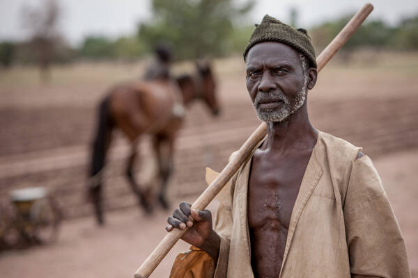 Portrait eines alten, dunkelhäutigen Mannes, der einen Stock über die Schulter gelegt hat. Im Hintergrund sieht man ein Pferd, das als Arbeitstier eingesetzt ist.