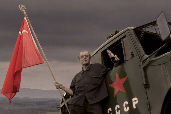 Ein schlampit Uniformierter Mann lehnt an einem sowietischen Militärfahrzeug mit einer sowietischen Fahne und hält eine CocaCola-Flasche in der hand.