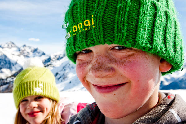 Close-up-Portrait eines sommersprossigen Buben mit grüner Planei-Strickhaube. Auf der Haube und im Gesicht sind Schneereste. hinter ihm ist eine Mädchen mit einer hellgrünen Haube. Beide sind umringt von den verschneiten Bergen rund um die Planai.