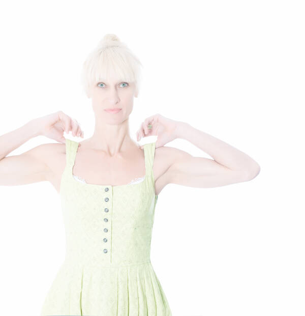 Highkey Aufnahme einer blonden Frau in gelbgrünem Trachtenkleid. Die beiden Daumen auf den Schultern unter den Trägern.