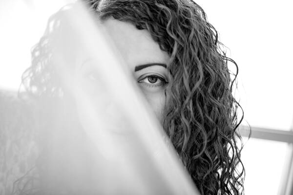 Portrait einer jungenFrau mit lockigem, langem Haar, schräg durchs Bild verdeckt ein Schleier den Blick auf ihr Gesicht, so dass nur ein Auge gut sichtbar ist. Schwarz-Weiß.