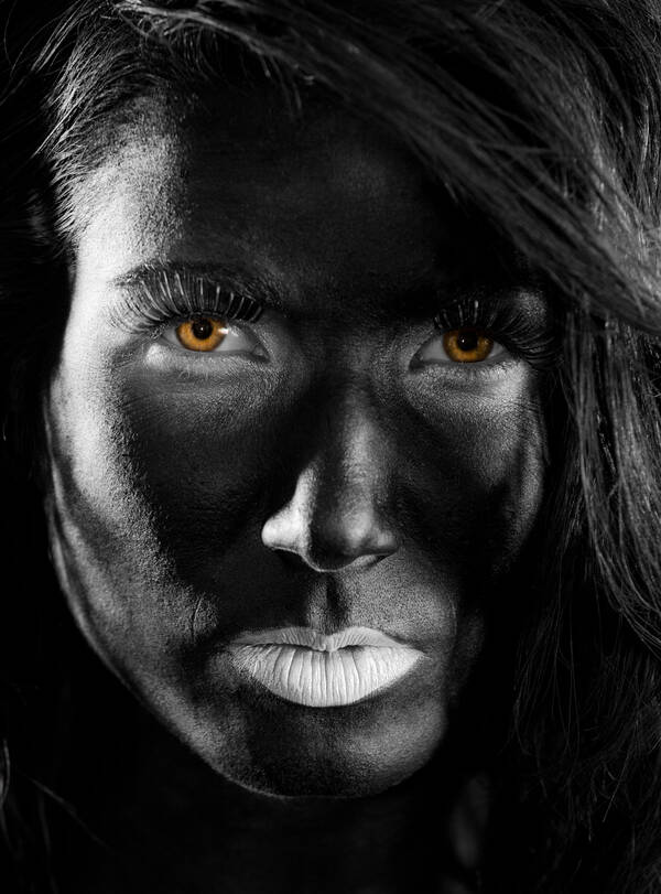 Close-up-Portrait einer Frau, deren Gesicht ganz dunkel geschminkt ist. Bis auf die leuchtend gelben Augen ist das Bild in Schwarz-Weiß.