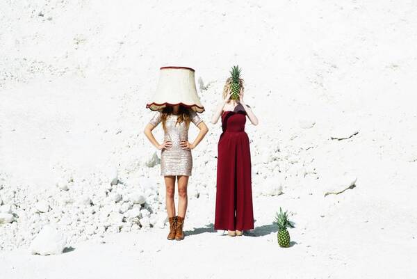 Zwei Frauen stehen in einem Steinbruch. Eine trägt einen Lampenschirm auf dem Kopf, die andere hält eine Ananas vor ihr Gesicht.