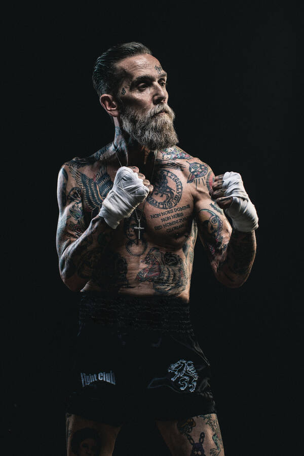 Ein älterer Mann mit Vollbart steht in Boxershorts mit bandagierten Händen wie im Boxkampf. Muskulös, stark tätowiert, ein Kreuz an einer Kette um den Hals. Low Key.