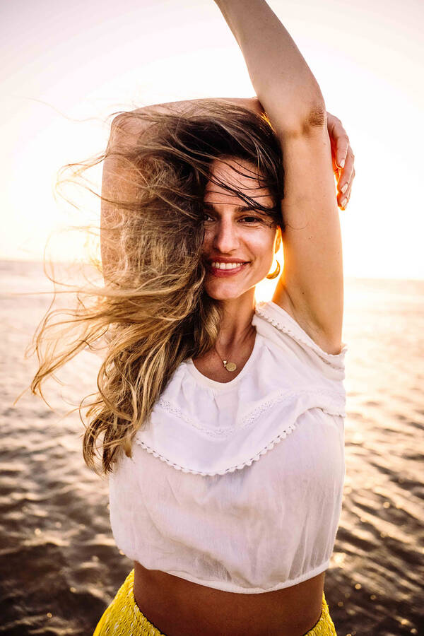 Eine Frau steht mit Windzerzausten Haaren vor einer Wasserfläche im Gegenlicht.