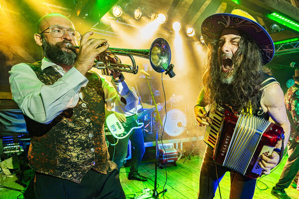 Musiker (Trompete, Ziehharmonika, E-Bass) auf einer Bühne in grüngelbem Licht.