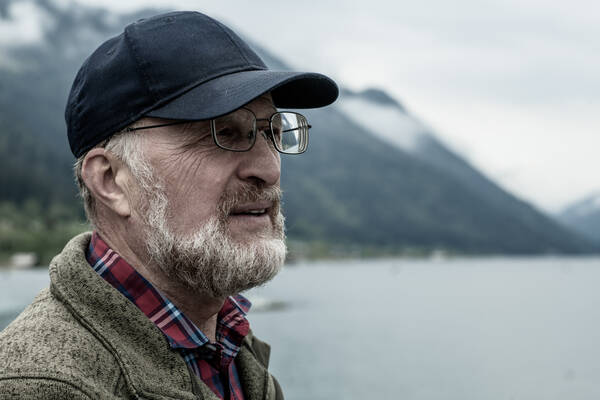 Ein bärtiger Mann mit Brille und Baseball-Kappe vor einem See in den Bergen.